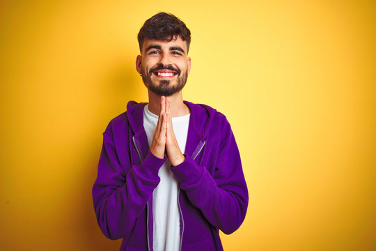 Young man with tattoo wearing sport purple sweatshirt over isolated yellow background praying with hands together asking for forgiveness smiling confident.