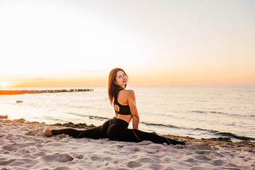 Fototapeta na wymiar Slim strong young woman in black practicing yoga doing the splits on sand beach close-up with copy space. Sunset, summer. Healhy lifestyle concept.