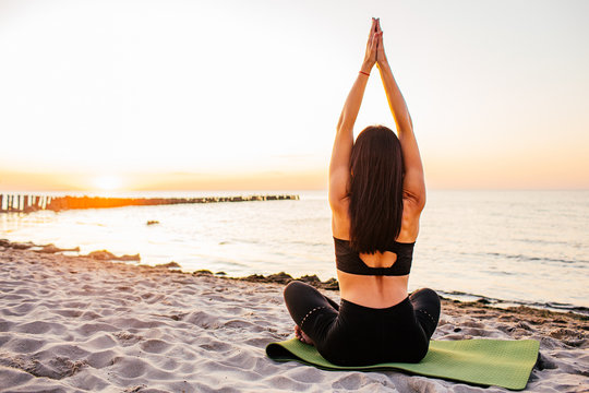 Young brunnete woman in a lotus pose with her hands up on the beach in a summer evening. Sunset. Meditation, concentration. Yoga practice.