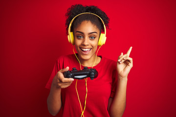 Afro woman playing video game using joystick and headphones over isolated red background very happy pointing with hand and finger to the side © Krakenimages.com