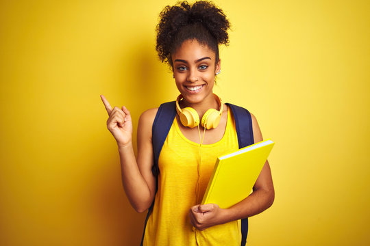 Afro woman using backpack and headphones holding notebook over isolated yellow background very happy pointing with hand and finger to the side