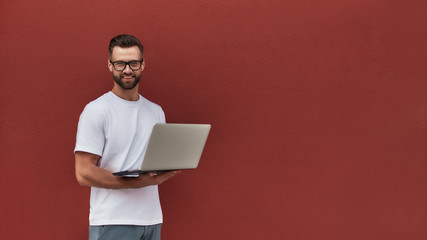 Happy blogger. Handsome young man in casual clothes and eyeglasses holding laptop and looking at camera with smile while standing against red wall outdoors