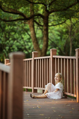 Fototapeta premium Adorable little girl with long blonde hair sitting on wooden bridge in park sunny summer day. Beautiful child. Happy smiling kid, childhood day, education concept