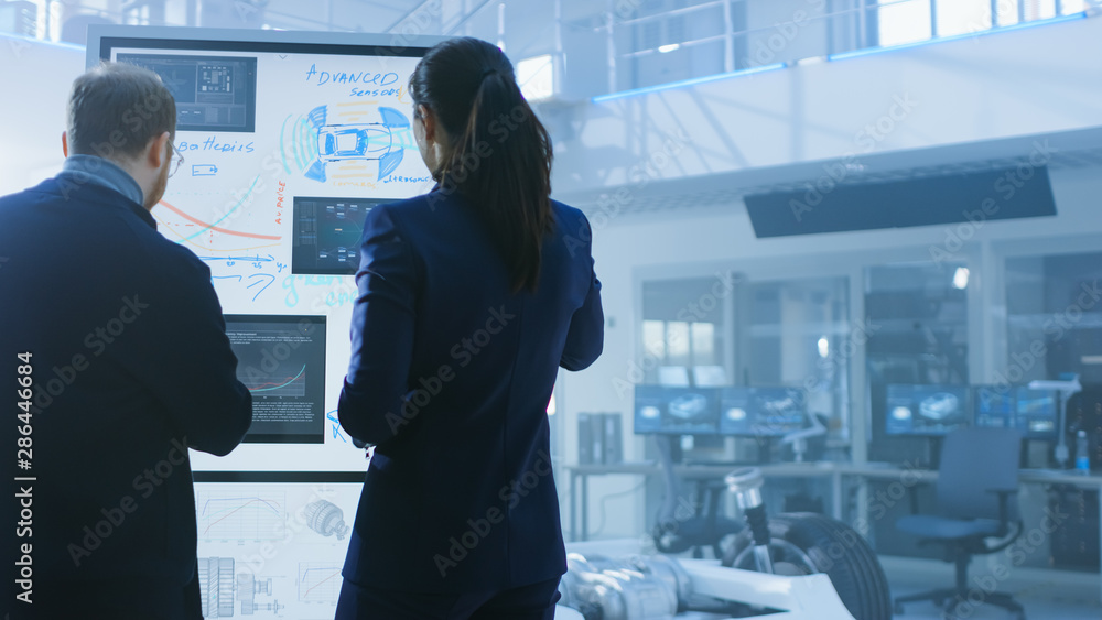 Poster Male and Female Design Engineers are Working and Discussing Something on a Interactive Whiteboard Next to an Electric Car Chassis Prototype. In High Tech Laboratory Facility with Vehicle Frame.