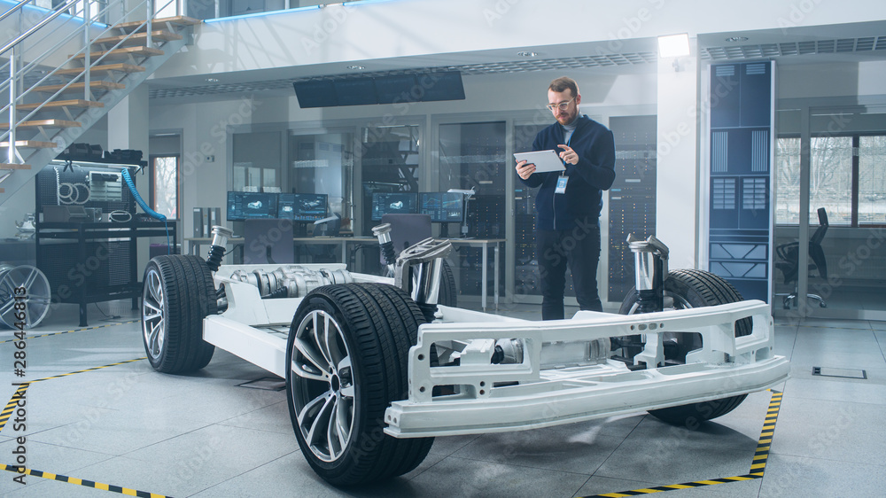 Poster Engineer with Glasses and Beard Scans Electric Car Chassis Prototype with Wheels, Batteries and Engine with an Augmented Reality Software on a Tablet Computer in a High Tech Development Laboratory.
