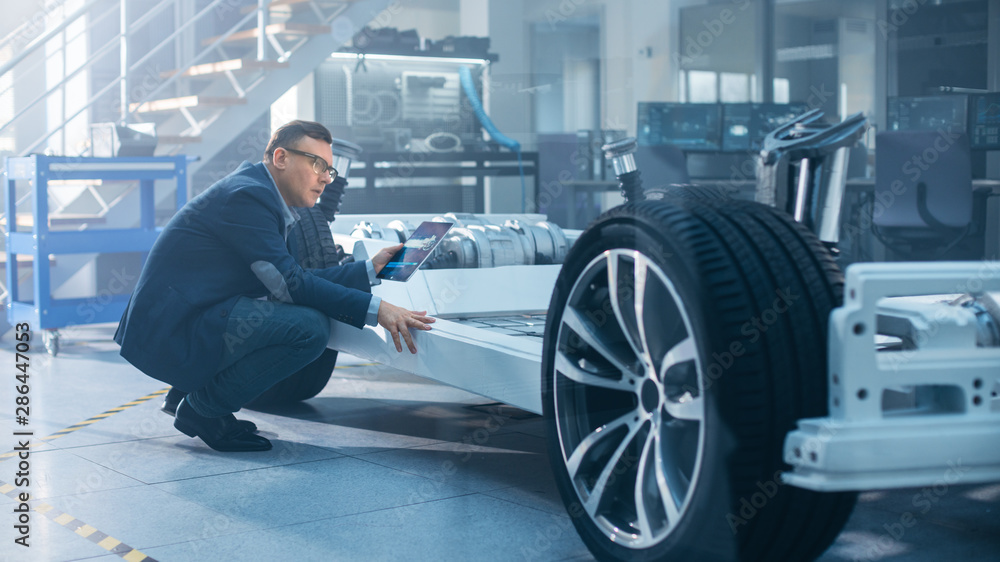Poster Engineer with Glasses Works on a Tablet Computer Next to an Electric Car Chassis Prototype with Wheels, Batteries and Engine in a High Tech Development Laboratory.