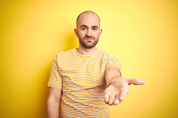 Young bald man with beard wearing casual striped t-shirt over yellow isolated background smiling cheerful offering palm hand giving assistance and acceptance.