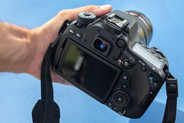 Close up of large black DSLR camera held in frame by a hand in front of light blue background.