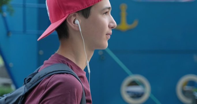 Teenager walking on the street and  listen to the music in earphones. Student in red baseball cap listens to the music in headphone, outdoors. Teenage boy in earphones goes outdoors. Urban scene.