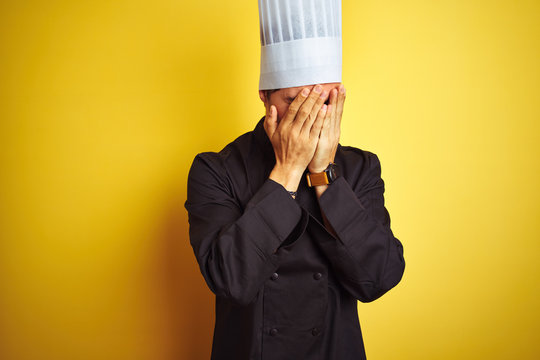 Young chef man wearing uniform and hat standing over isolated yellow background with sad expression covering face with hands while crying. Depression concept.