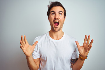 Young handsome man with tattoo wearing casual t-shirt over isolated white background crazy and mad shouting and yelling with aggressive expression and arms raised. Frustration concept.
