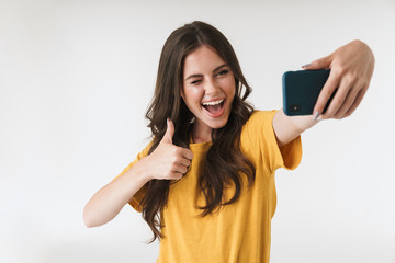 Fototapeta premium Image of young brunette woman laughing and showing thumb up while taking selfie photo on cellphone