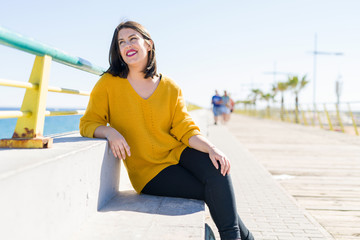 Beautiful young woman sitting on a promenade by the sea smiling cheerful enjoying sunlight relaxing on sunny day