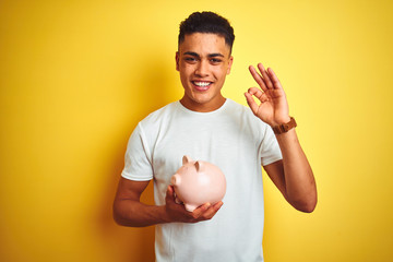 Fototapeta na wymiar Young brazilian man holding piggy bank standing over isolated yellow background doing ok sign with fingers, excellent symbol