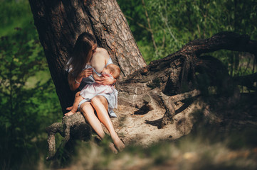 Happy mother breastfeeding her baby under a tall tree in a summer forest at noon.