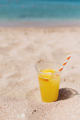 Glass of refreshing lemonade with a tube on a sand beach with blue ocean background. Trip and summer relax concept. Copy space.
