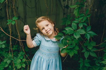 Cute little girl is standing in garden between green leaves of grapes and she looking to camera.