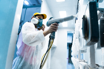 Powder coating of metal parts. A woman in a protective suit sprays white powder paint from a gun on...