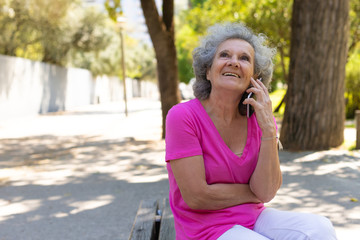 Happy serene old lady talking on cell in park. Joyful senior grey haired woman in casual sitting on bench and speaking on mobile phone. Nice phone talk concept