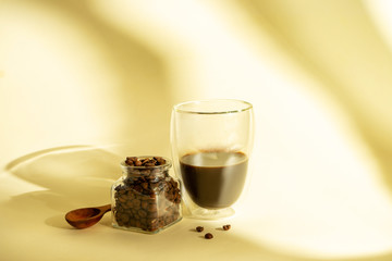 Morning coffee concept. Stylish poster for coffee. double-walled glass with poured coffee and grain on yellow background with shadows. Copy space. Flat lay food