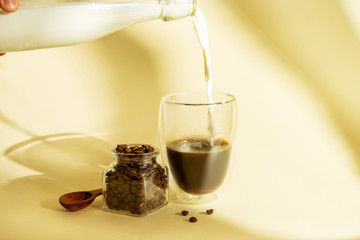 Morning coffee concept. Stylish poster for coffee. Pouring milk from bottle into double-walled glass with poured coffee and grain on yellow background with shadows. Copy space. Flat lay food