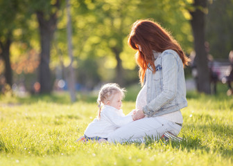 Happy pregnant mother and daughter in the autumn park. Beauty nature scene with family outdoor lifestyle. Happy woman and girl resting together on green grass, having fun outdoor