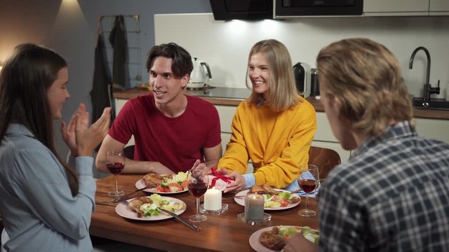 Group of four joyful friends, two couples, having festive dinner in domestic kitchen. Young woman presenting gift box and wishing all the best to girlfriend at birthday party