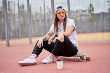 Fototapeta premium Image of athletic brunette girl in sunglasses sitting on skateboard on playground on summer day