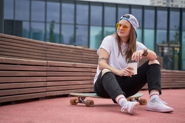 Photo of sports brunette in sunglasses in her hands sitting on skateboard on sports field near...