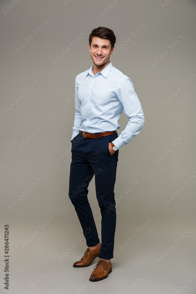 Poster image of happy young man wearing formal clothes smiling at camera with hands in pockets