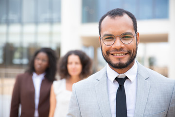 Cheerful African American businessman. Portrait of handsome young businessman smiling at camera while female colleagues standing behind. Leadership concept