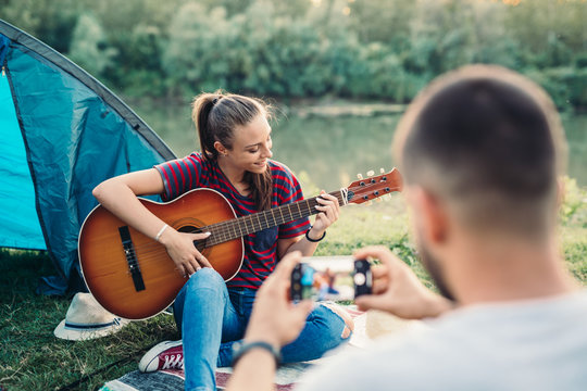 woman playing guitar while her boyfriend taking picture of her. camping by the lake