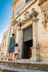 Fototapeta na wymiar A mother and her child climb the steps and enter the church of Cavallino, Lecce, Puglia, Salento, Italy. In baroque style. Wooden portal with statues on the sides, onthe facade.