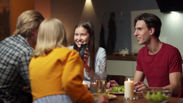 Zoom out shot of group of four friends, two young couples, having dinner at home together. Young woman showing picture of her boyfriend on smartphone screen, friends laughing