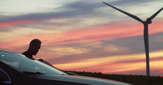 Man near car walking in the countryside lanes watching large wind turbines spinning in amazing pink sky sunset. Efficient clear energy wind power station.