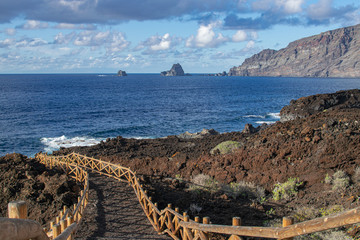 Volcanic pathway with wooden railing, with Roques de Salmor, Atlantic ocean, blue sky and clouds...