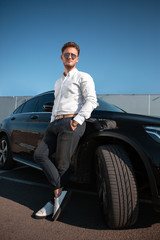 Fototapeta na wymiar Young successful businessman in a white shirt and sun glasses standing near his luxary black car.