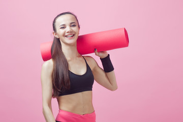 A brunette girl, attractive, is standing, holding a yoga mat over the shoulders.