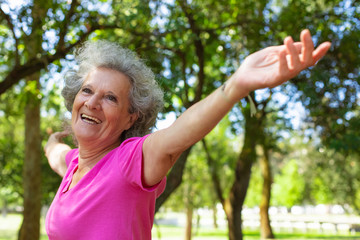 Happy excited old lady enjoying yoga class outdoors. Senior grey haired woman in casual standing in park, outstretching hands and laughing. Age and outdoor fitness concept