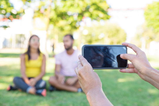 Person with phone taking picture of couple relaxing in park. Mobile phone in human hands and man and woman sitting on grass in park and chatting. Photographing on smartphone concept