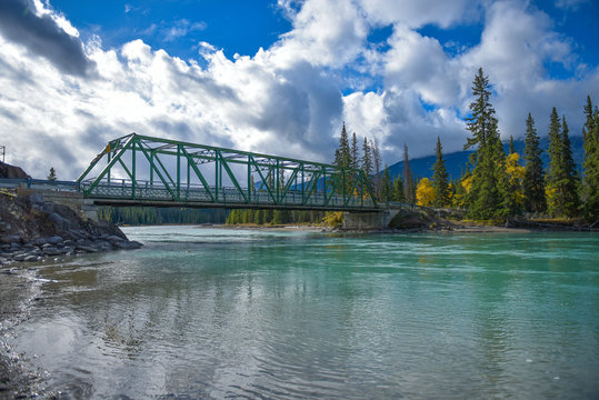 rio color turquesa que pasa bajo un puente de metal en un bosque rodeado por monta&ntilde;as nubes frises y un cielo azul en el parque nacional Jasper Canad&aacute;