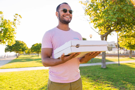Happy Latin male courier carrying pizza in park. Handsome young man standing on grass, holding pizza boxes, looking away and smiling. Food order concept