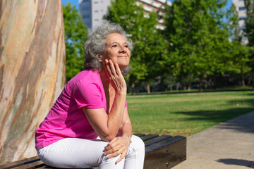 Joyful old lady waiting someone in city park. Senior grey haired woman in casual sitting on bench outdoors, leaning chin on hand and looking into distance. City park concept