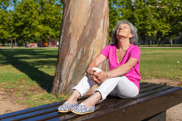 Peaceful calm old lady enjoying leisure in park. Senior grey haired woman in casual sitting and placing feet on bench outdoors, turning face up with closed eyes. Leisure in park concept