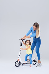 Young woman and little girl standing on scooter and skateboard. Mom teaches daughter to ride, have a fun and joyfully looking in camera over grey background