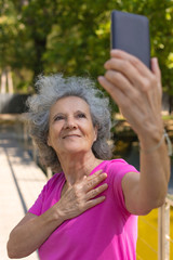 Cheerful delighted old lady chatting through video call. Senior grey haired woman in casual talking on smartphone while walking in park. Video chat concept