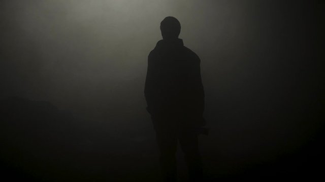 Black silhouette of man standing in smoky dark. Stock footage. Mysterious silhouette of young man stands in darkness lit only by dim light in haze