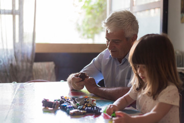 Caucasian grandfather and toddler granddaughter playing with toy cars in indoor location with natural light. Copy space on the left.