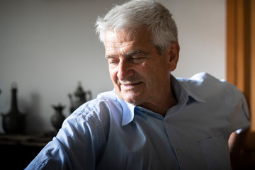 Older caucasian man with grey hair, wearing a blue shirt, smiling and looking away in indoor location with natural light.