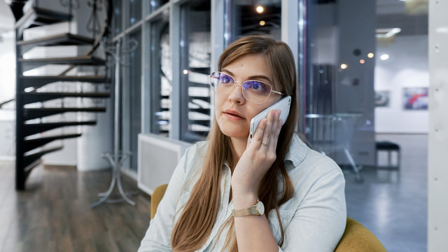 Sharing Shock Business News. Attractive Business Woman Talking On The Mobile Phone Seriously While Sitting At Her Working Place In Office And Working With Documents